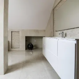 Jasna łazienka na poddaszu z beżowymi płytkami, białą szafką pod umywalką i dekoracyjną czarną wazą w niszy.