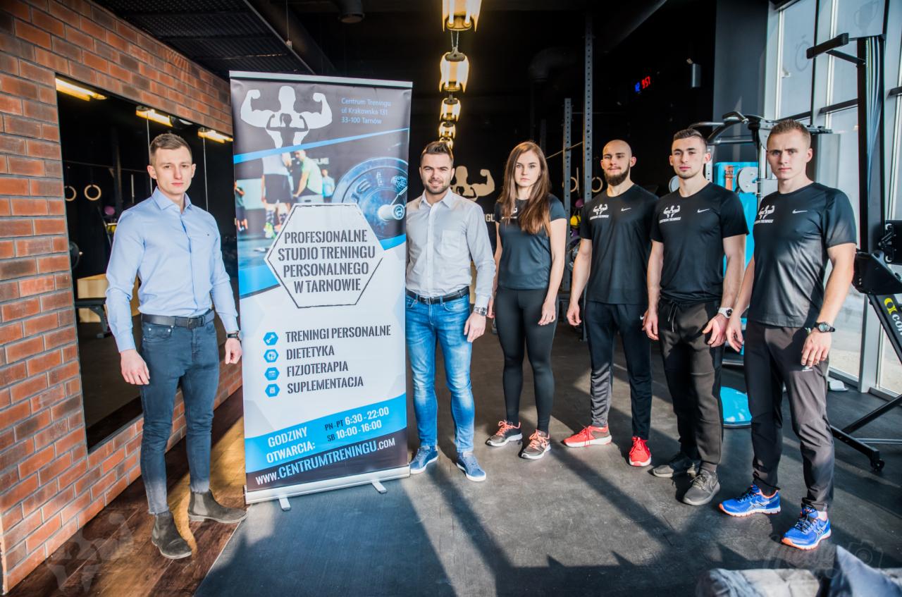 Zespół studia treningu personalnego w Tarnowie prezentuje się na tle ściany z cegły i baneru reklamowego z ofertą treningów personalnych, dietetyki, fizjoterapii i suplementacji.