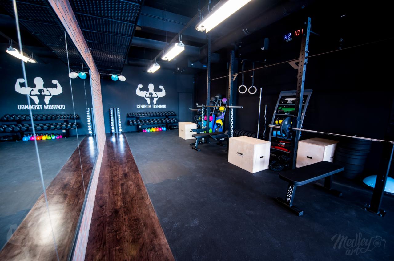 Wnętrze nowoczesnej siłowni z czarnymi ścianami, wyposażonej w stojaki na hantle, kettlebells, sztangi, drążki do podciągania, skrzynie do ćwiczeń i lustro z logo centrum treningowego.