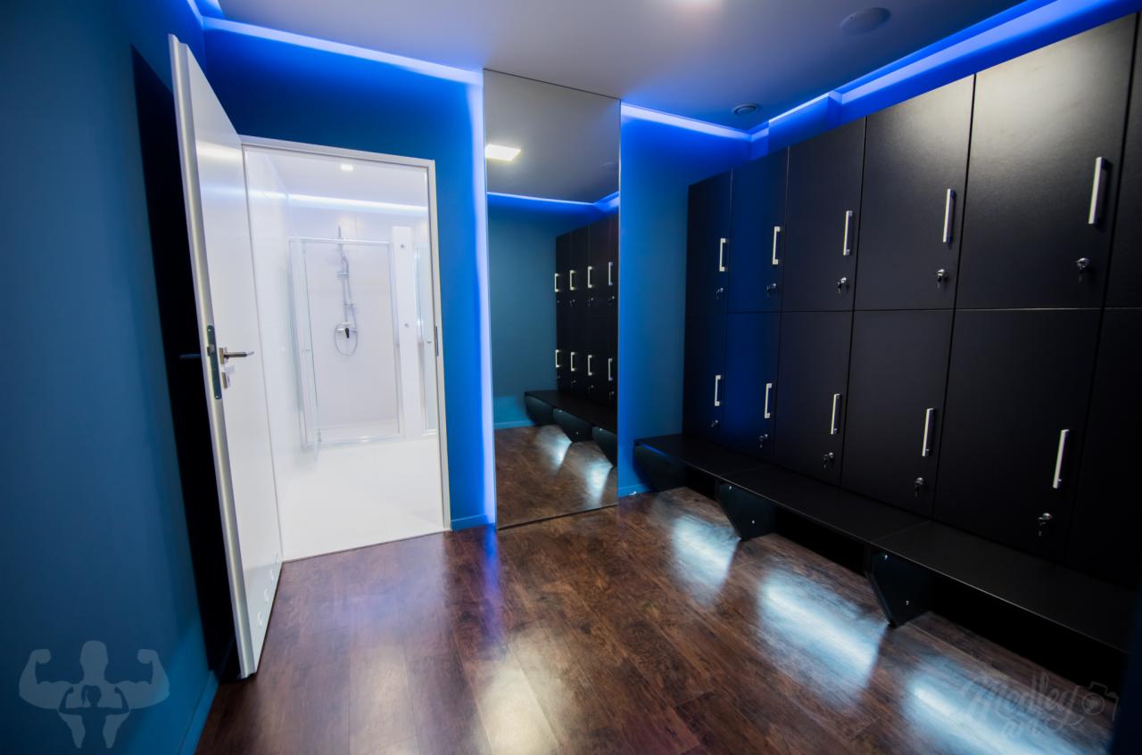 Nowoczesna szatnia z czarnymi szafkami, drewnianą podłogą i niebieskim oświetleniem LED, widoczny prysznic z otwartymi drzwiami.