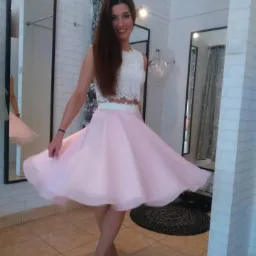 Uśmiechnięta kobieta w przymierzalni, prezentująca zwiewną, różową spódnicę z tiulu i koronkowy top, obracając się lekko, z widocznymi trzema lustrami i białą cegłą na ścianach.