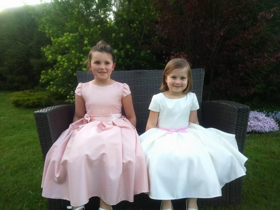 Dwie uśmiechnięte dziewczynki w eleganckich sukienkach siedzą na wiklinowym krześle ogrodowym na tle zieleni.