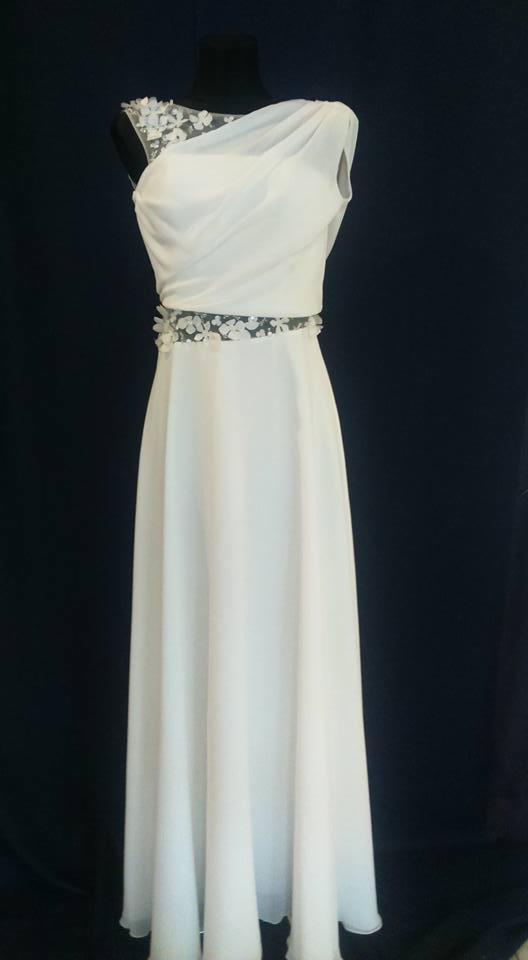 Elegancka, długa, biała suknia z asymetrycznym dekoltem, ozdobiona kwiatowymi aplikacjami na ramieniu i w pasie, prezentowana na manekinie na ciemnym tle.