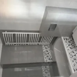 Wykończona łazienka z szarymi płytkami o geometrycznym wzorze, białą grzejnikiem drabinkowym i podtynkową spłuczką WC.