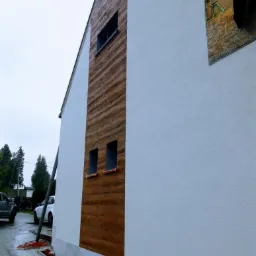 Fragment elewacji domu z częściowo wykończoną strukturą: biały tynk obok paneli imitujących drewno, widoczne okna i przygotowania do dalszych prac na zewnątrz budynku.