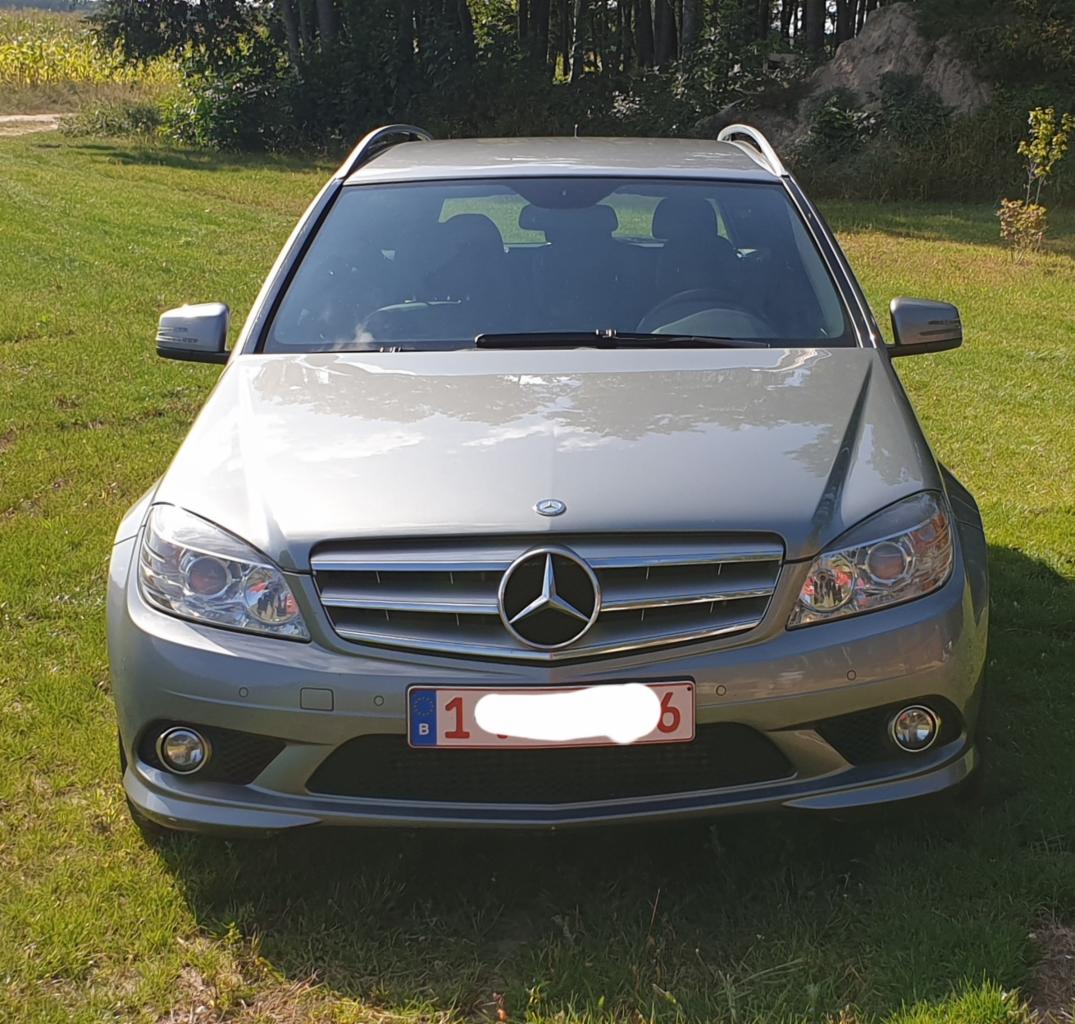 Szary Mercedes C-klasa kombi z belgijską tablicą rejestracyjną, zaparkowany na trawie, z widocznymi reflektorami i logo marki. Przód pojazdu.