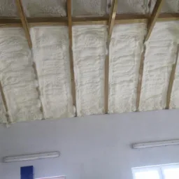 Poddasze ocieplone białą pianą natryskową, widoczne drewniane krokwie i fragment ściany z lampą fluorescencyjną.