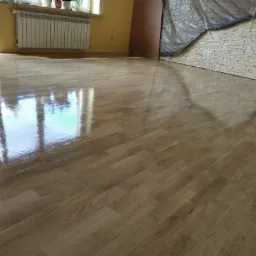 Świeżo polakierowana, błyszcząca podłoga parkietowa z widocznym odbiciem okna, zabezpieczona folią ściana w tle.