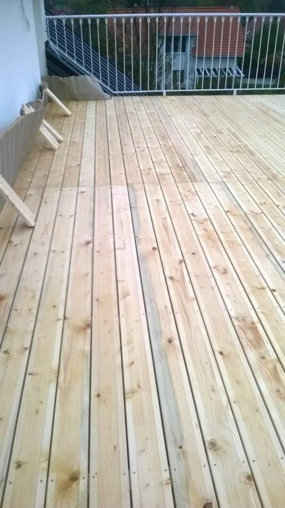 Deski podłogowe z jasnego drewna na balkonie, widoczne białe barierki i fragment budynku w tle.