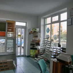 Wnętrze biura z ekspozycją różnorodnych żaluzji wewnętrznych, próbek materiałów i profili okiennych, z widokiem na ulicę przez okna PCV.