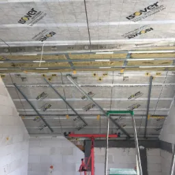 Zabudowa poddasza: widok na sufit z izolacją wełną mineralną, metalową konstrukcją nośną i folią paroizolacyjną Isover Vario KM Duplex UV, częściowo zamontowaną, widoczny podnośnik do płyt...