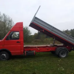 Czerwona wywrotka z podniesioną skrzynią ładunkową, widok z boku na tle zielonego trawnika i drzew.