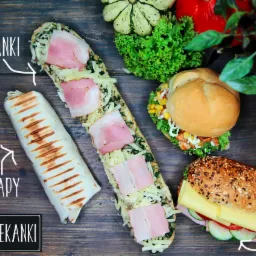 Widok z góry na drewniany stół z różnorodnym jedzeniem: wrap, zapiekanka z szynką, kanapki z warzywami i serem, dynie ozdobne i zielenina