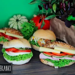 Cztery apetyczne kanapki na drewnianym blacie, z różnymi dodatkami: sałata, pomidor, ogórek, ser, szynka, sos, w tle dekoracja z warzyw i ziół w papierowej torbie, logo 'MEGA ZAPIEKANKI' w dolnej...