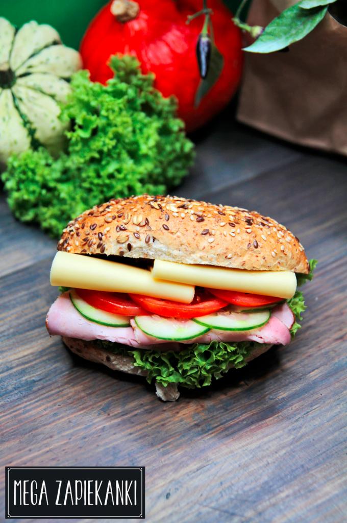 Kanapka z szynką, serem, pomidorem i ogórkiem na ziarnistym pieczywie, otoczona warzywami i dyniami na drewnianym stole, z logo 'MEGA ZAPIEKANKI' w rogu.