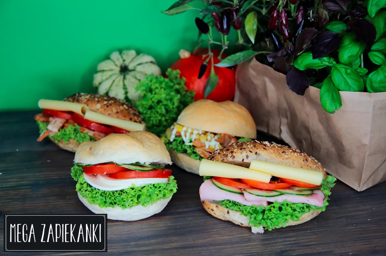 Cztery apetyczne kanapki na drewnianym blacie, z różnymi dodatkami: sałata, pomidor, ogórek, ser, szynka, sos, w tle dekoracja z warzyw i ziół w papierowej torbie, logo 'MEGA ZAPIEKANKI' w dolnej...