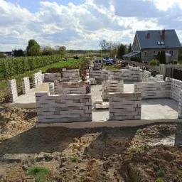 Wznoszone ściany parteru domu murowanego z betonu komórkowego na fundamencie, widok na sad owocowy w tle.