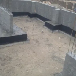 Betonowe ściany fundamentowe z hydroizolacją bitumiczną w gruncie, widoczne zbrojenia pionowe, w tle sterta ziemi i drzewa bez liści.