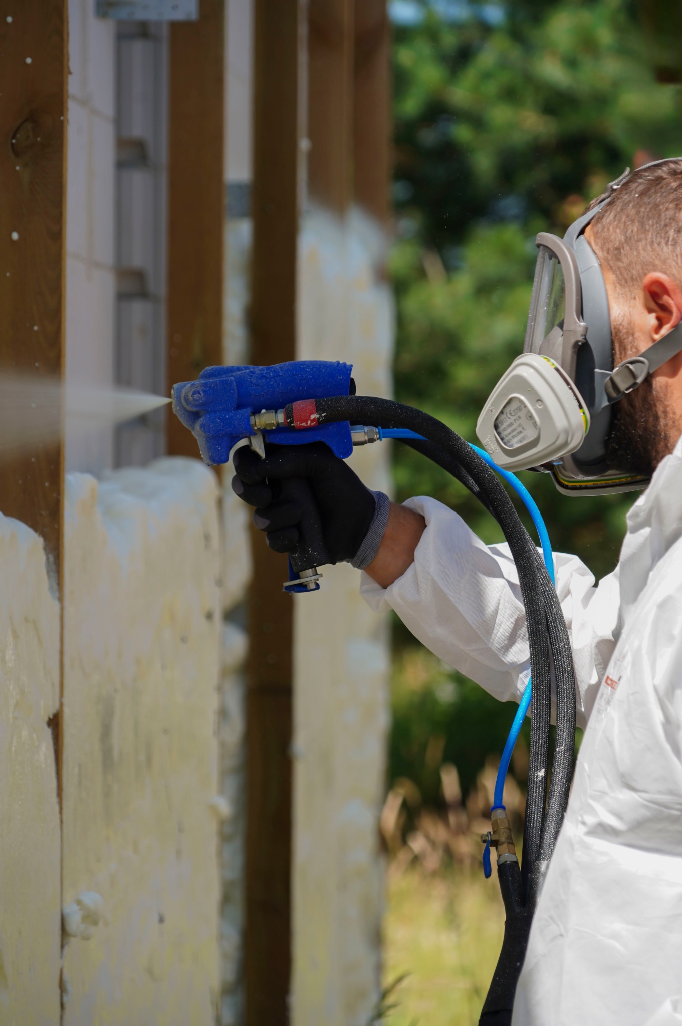 Pracownik w kombinezonie i masce ochronnej natryskuje piankę PUR na drewnianą konstrukcję ściany, tworząc izolację termiczną.