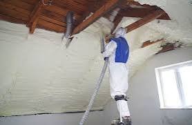 Pracownik w kombinezonie ochronnym aplikuje piankę izolacyjną na poddaszu w Stanisławicach. Widoczne drewniane elementy konstrukcji dachu i fragment okna.