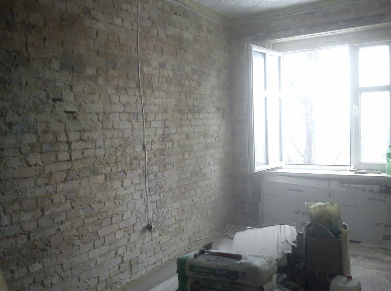Wnętrze pokoju w trakcie remontu: odsłonięta cegła na ścianie, nowe okno PCV, worki z gipsem i narzędzia na podłodze, przygotowania do dalszych prac wykończeniowych.