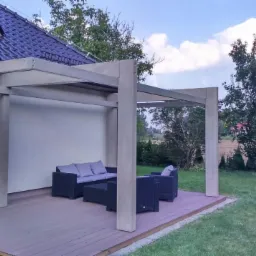 Szara pergola z płaskim dachem i kwadratowymi słupami na tarasie z desek kompozytowych, z widocznym zestawem mebli ogrodowych w kolorze antracytowym, przylegająca do domu z ciemnym dachem i ogrodem...