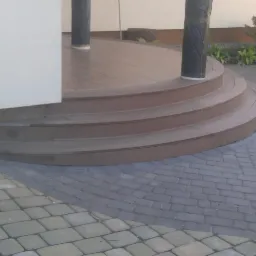 Półokrągłe schody prowadzące na taras z desek, obok ułożona kostka brukowa, widoczne słupy owinięte folią ochronną.