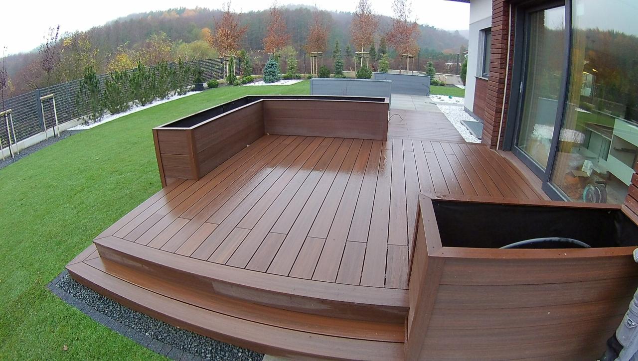 Nowoczesny taras z desek kompozytowych w kolorze brązu, z drewnianymi donicami, schodami i widokiem na zadbany ogród z zielonym trawnikiem oraz drzewami w tle.