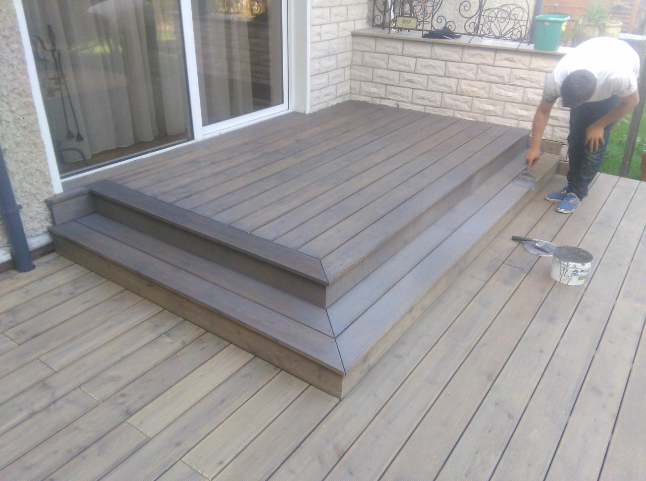 Malowanie drewnianego tarasu z trzema stopniami, widoczna puszka z farbą i pędzel, osoba w trakcie pracy, jasne drewno poddane renowacji.