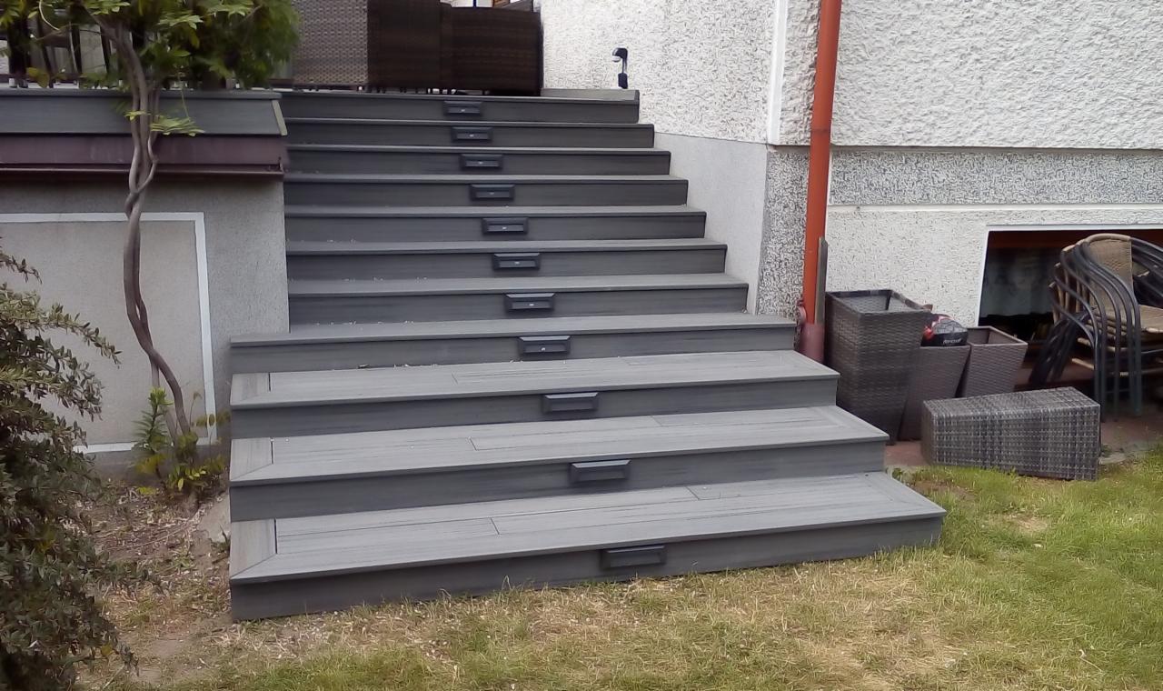 Schody zewnętrzne z desek kompozytowych w kolorze szarym, prowadzące do domu. Na każdym stopniu zamontowane małe lampki oświetleniowe. Obok schodów widoczne donice i meble ogrodowe.