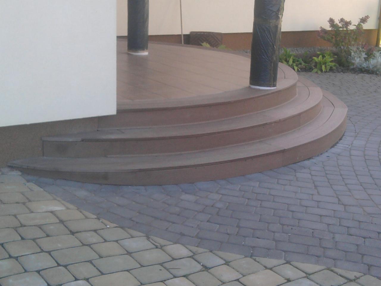 Półokrągłe schody prowadzące na taras z desek, obok ułożona kostka brukowa, widoczne słupy owinięte folią ochronną.
