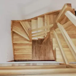 Widok z góry na kręte, jasne schody z drewna w Dąbrowie, z balustradą z prostych szczebli i podłogą wyłożoną panelami imitującymi drewno.