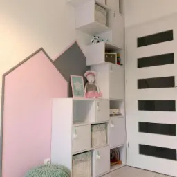 Białe modułowe regały dziecięce z kwadratowymi schowkami, niektóre z wiklinowymi koszami, ustawione w narożniku pokoju z dekoracją ścienną w kształcie domku i turkusową pufą.