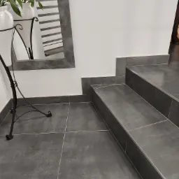 Szare płytki podłogowe i schodowe z delikatną fugą, lustro z odbiciem rolet i rośliny w białej doniczce na metalowym stojaku, białe ściany.