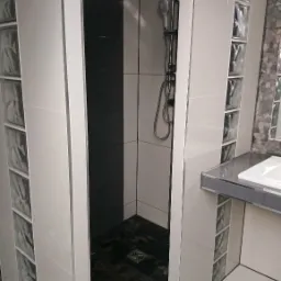 Nowoczesna kabina prysznicowa walk-in z czarnymi płytkami na podłodze i szklanymi luksferami w ścianie, kwadratowa deszczownica i bateria prysznicowa w kolorze chromu.