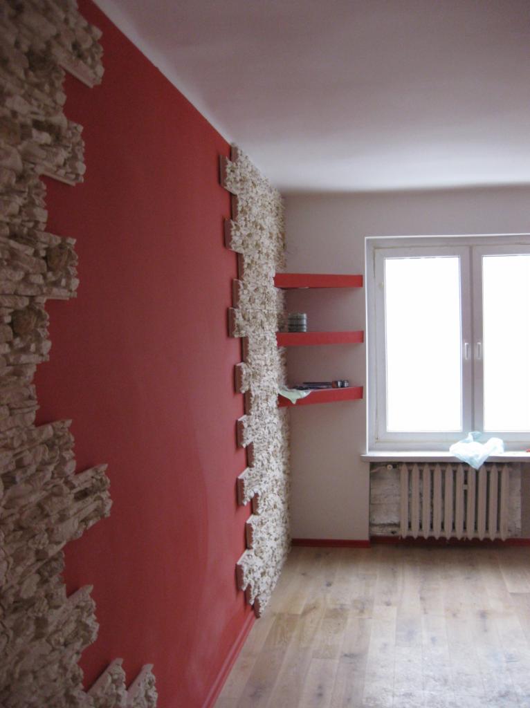 Czerwona ściana z dekoracyjnym kamieniem w narożniku, fragment ściany wykończony kamieniem dekoracyjnym, czerwone półki wiszące w pokoju z drewnianą podłogą i oknem.