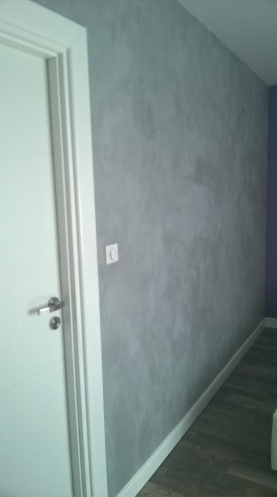 Ściana pokryta dekoracyjną szarą farbą strukturalną, widoczny fragment białych drzwi z chromowaną klamką oraz biała listwa przypodłogowa.