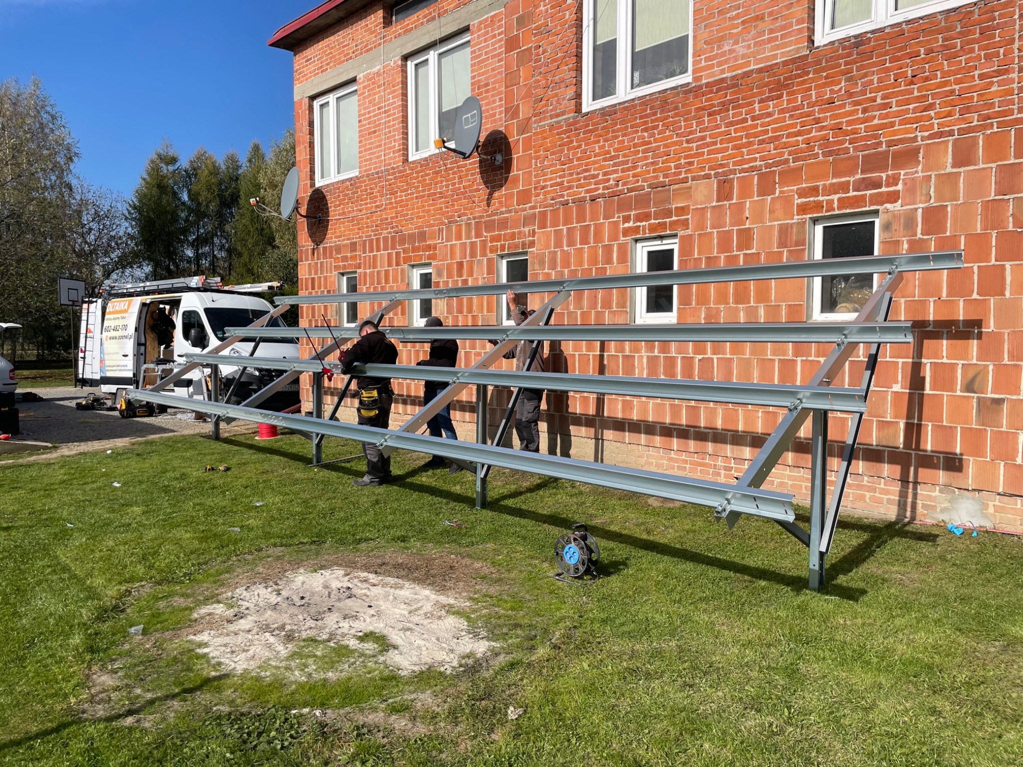 Montaż stalowej konstrukcji wsporczej pod panele słoneczne na trawniku przed budynkiem z czerwonej cegły; widoczni trzej monterzy i samochód dostawczy z logo firmy.