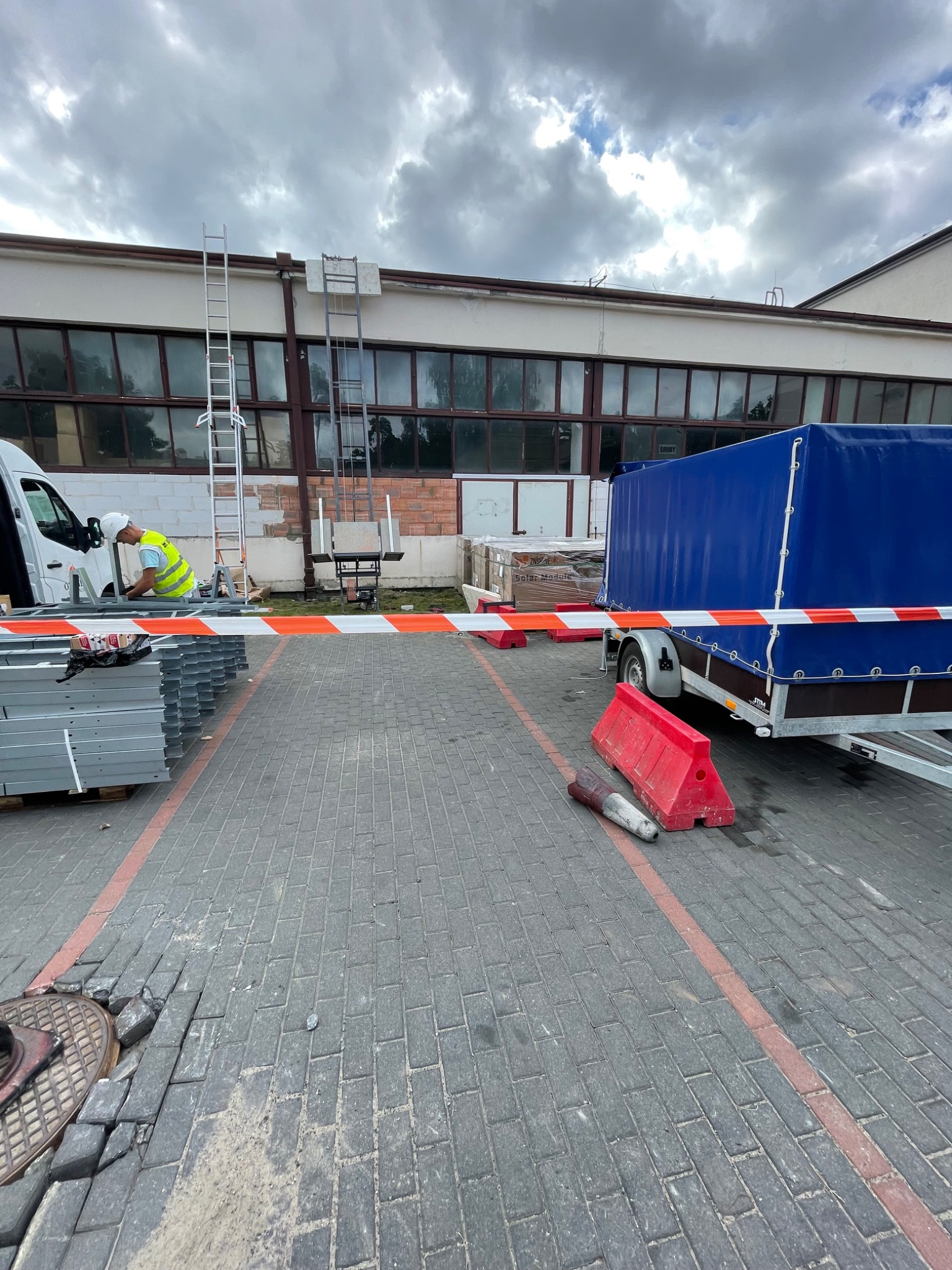 Pracownik w kamizelce i kasku przy samochodzie dostawczym, przygotowuje elementy konstrukcyjne, być może do montażu paneli fotowoltaicznych. W tle drabina oparta o budynek i przyczepa z niebieską...