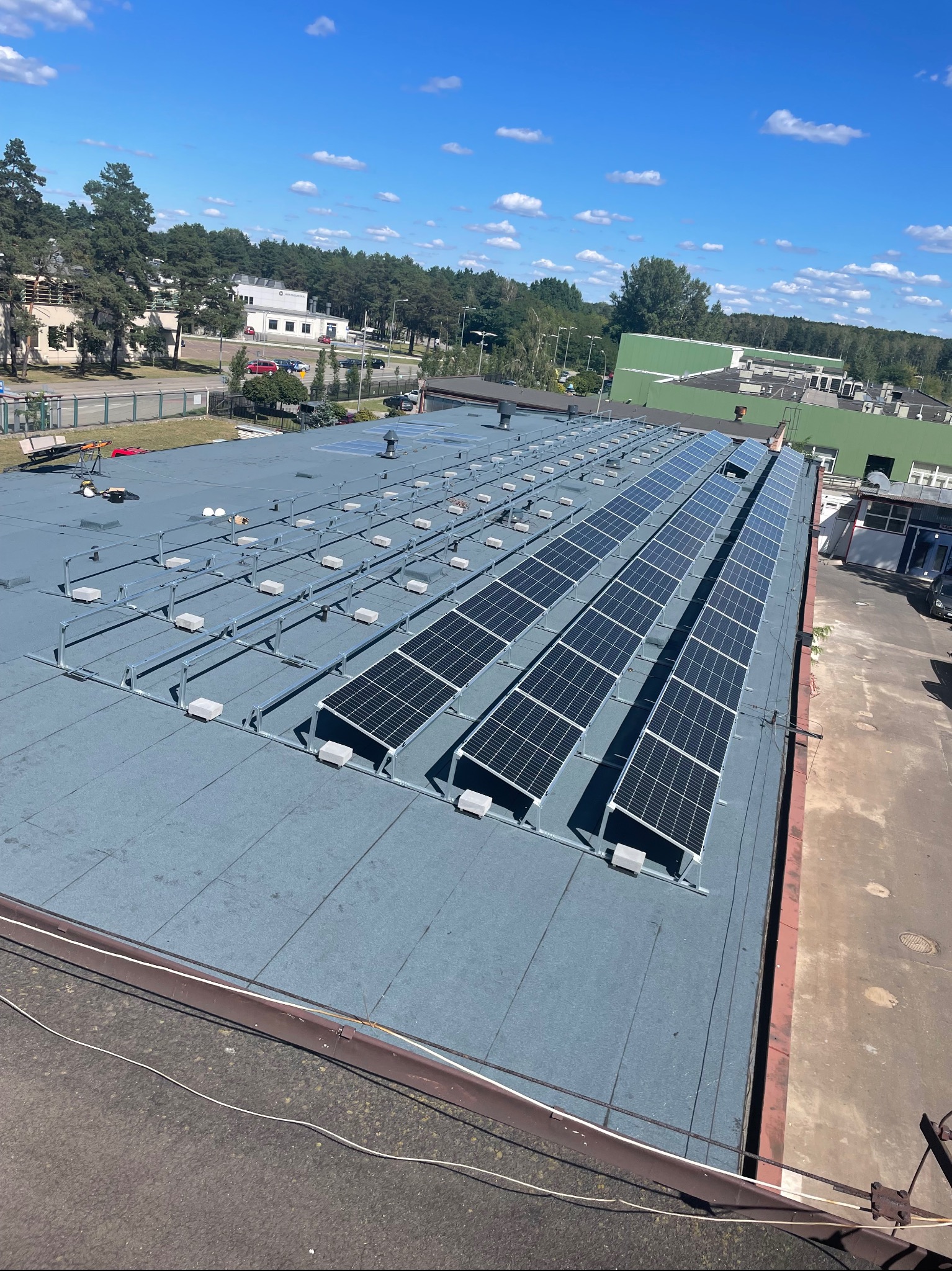 Panele słoneczne zamontowane na płaskim dachu budynku przemysłowego, widoczne elementy konstrukcyjne i okablowanie, w tle drzewa i błękitne niebo z chmurami.