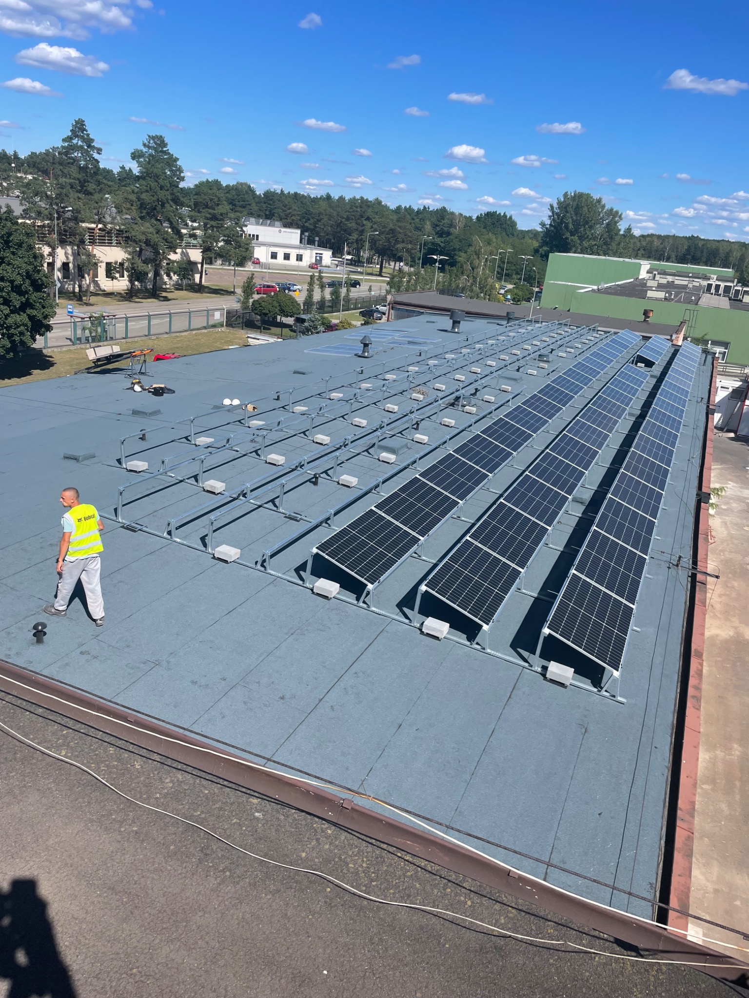 Montaż paneli słonecznych na dachu budynku przemysłowego, pracownik w kamizelce odblaskowej na tle rzędów paneli i konstrukcji wsporczych.