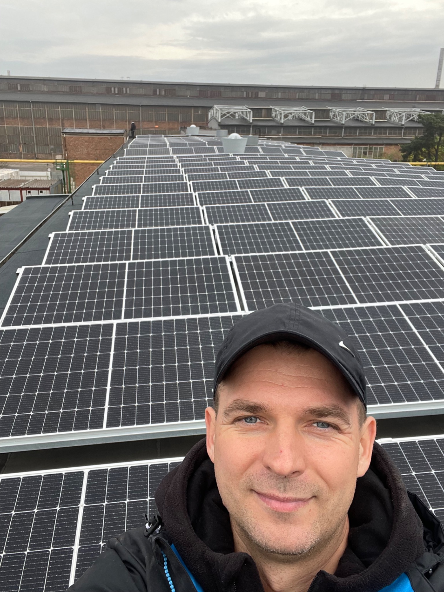 Selfie mężczyzny na dachu pokrytym panelami słonecznymi, w tle widoczny budynek przemysłowy i pochmurne niebo.