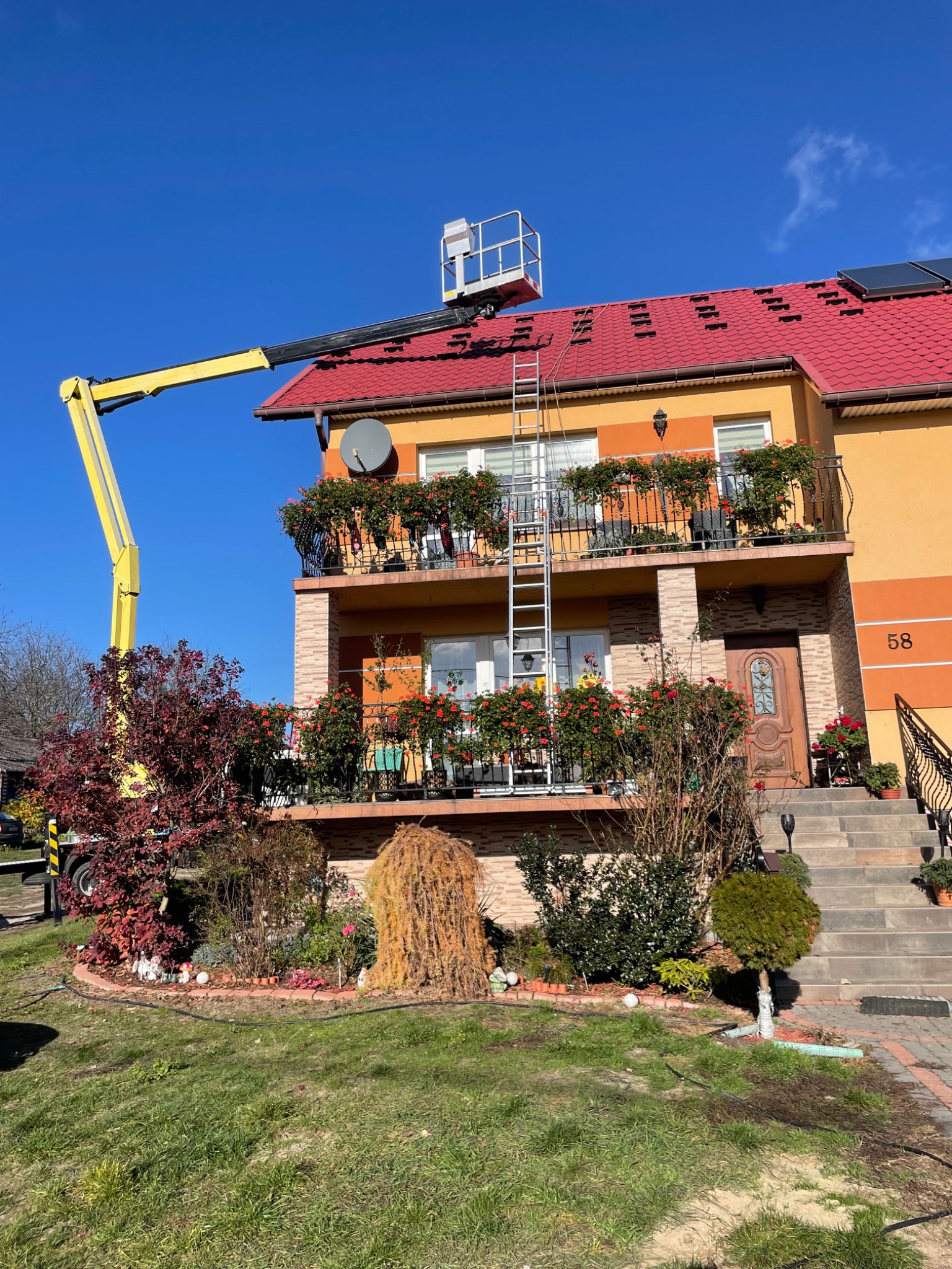 Żółty podnośnik koszowy na tle dwupiętrowego domu z czerwonym dachem i panelami słonecznymi, drabina oparta o dach, balkony z kwiatami, zadbany ogród.