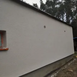 Świeżo otynkowana elewacja budynku z widocznym oknem zabezpieczonym folią i dwoma otworami na instalacje, na tle lasu.