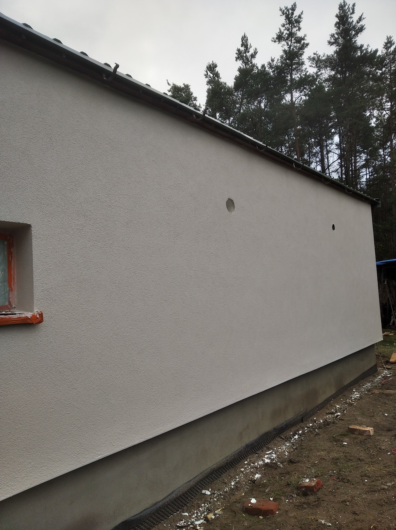 Świeżo otynkowana elewacja budynku z widocznym oknem zabezpieczonym folią i dwoma otworami na instalacje, na tle lasu.
