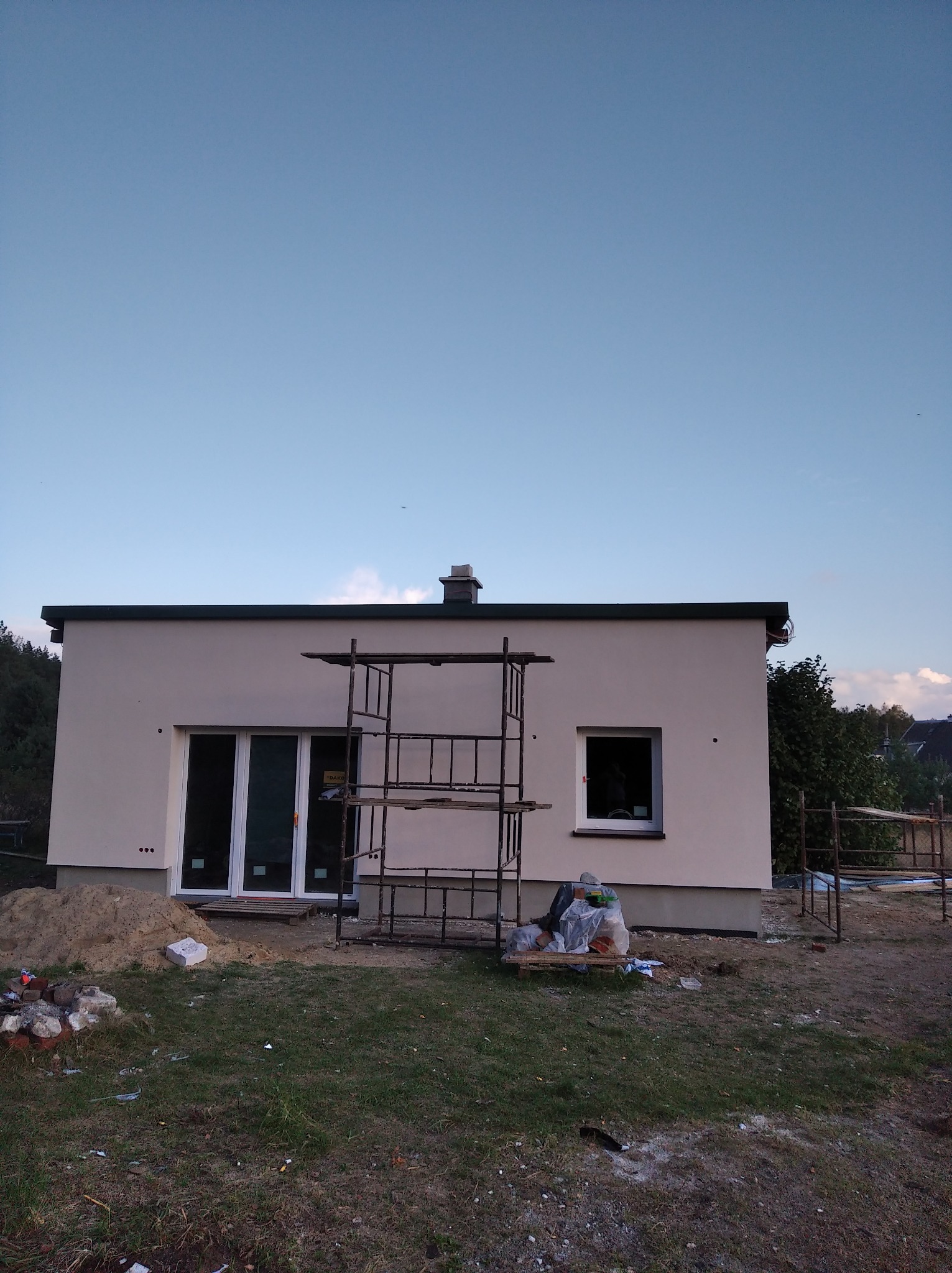 Nowo otynkowany dom jednorodzinny z zamontowanymi oknami i drzwiami balkonowymi, otoczony rusztowaniem i stertą piasku.