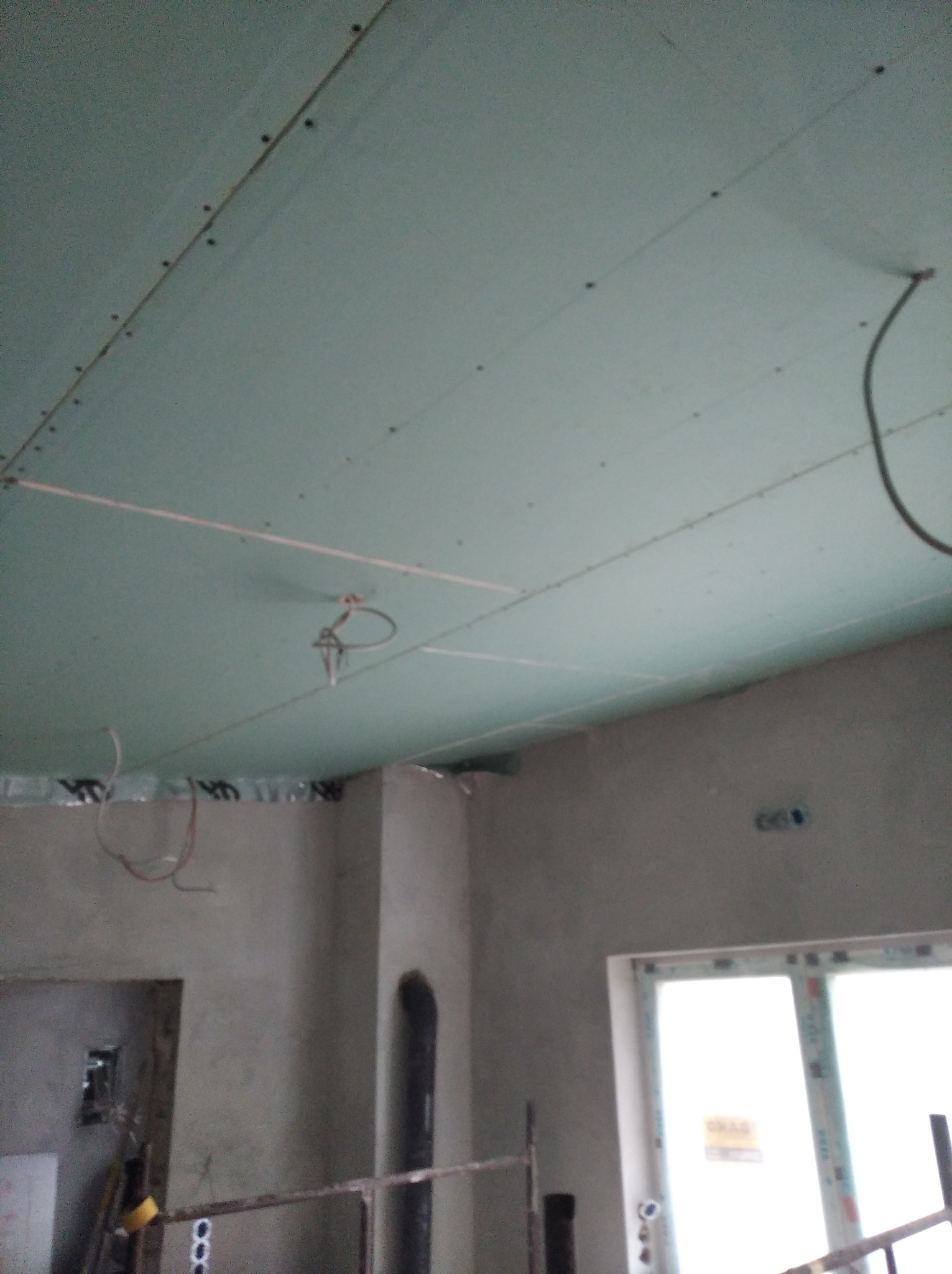 Niedokończony sufit podwieszany z płyt kartonowo-gipsowych, widoczne kable instalacji elektrycznej, fragment ściany z surowym tynkiem i okno z PCV.
