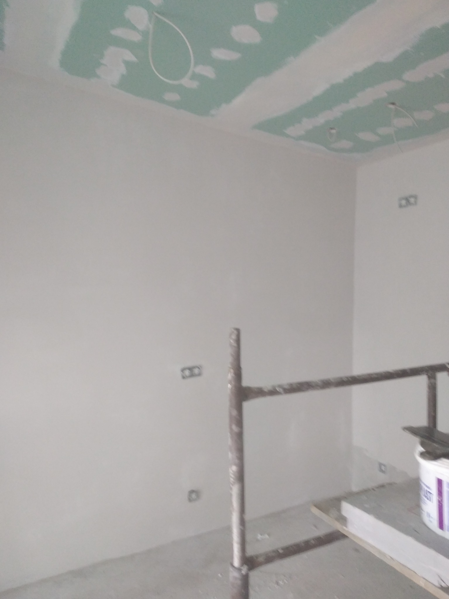 Świeżo zagruntowane ściany w szarym odcieniu, częściowo wykończony sufit z płyt gipsowo-kartonowych z widocznymi przewodami elektrycznymi, fragment rusztowania z wiadrem farby w rogu pomieszczenia.