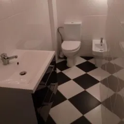 Biała łazienka z czarno-białą szachownicą na podłodze, widok na umywalkę z szafką, toaletę i bidet.