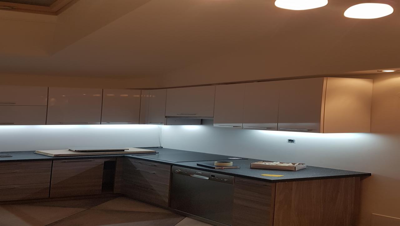 Nowoczesna kuchnia z białymi szafkami wiszącymi podświetlonymi ledami i drewnopodobnymi szafkami dolnymi, blat z ciemnego kamienia, widoczne akcesoria montażowe i narzędzia, prawdopodobnie...